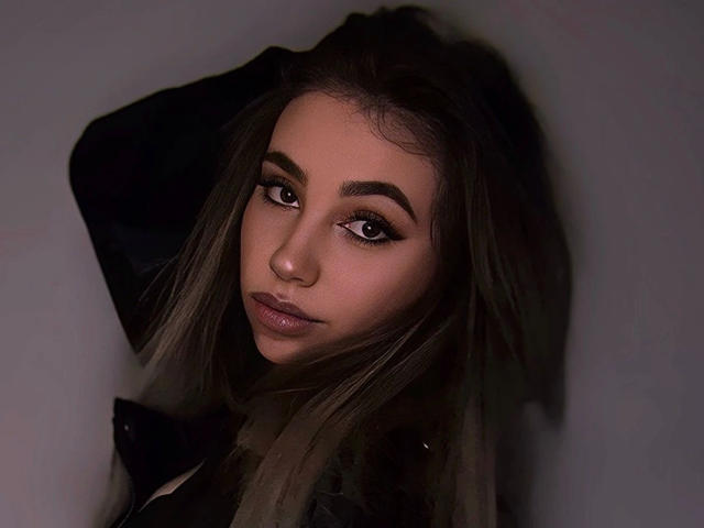 LilyLiebe-hot with dark eyes, thick eyebrows, long brown hair, and a serious look, wearing a black jacket, arms raised, in a dimly lit, dark-toned photo.