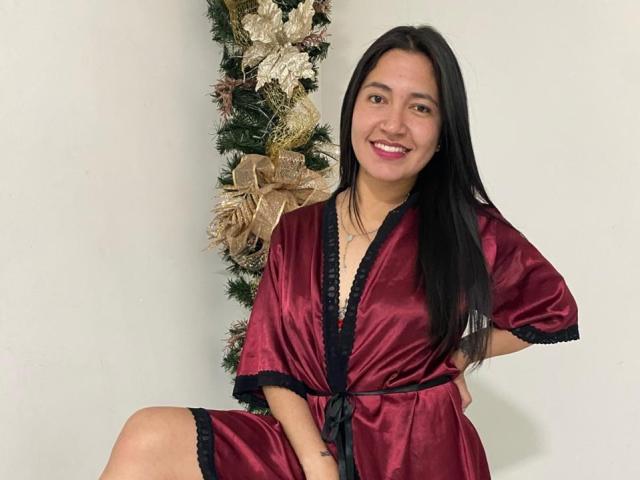 ValentinaHottest, a young woman with long black hair, sits in a red satin robe with black lace trim. She smiles at the camera, showing white teeth. Her right hand rests on her hip. A Christmas wreath with gold ribbons is in the background. Her skin is light brown, and she has a small tattoo on her right wrist. The robe's fabric is shiny and reflects light. She's in a simple, white-walled room.
