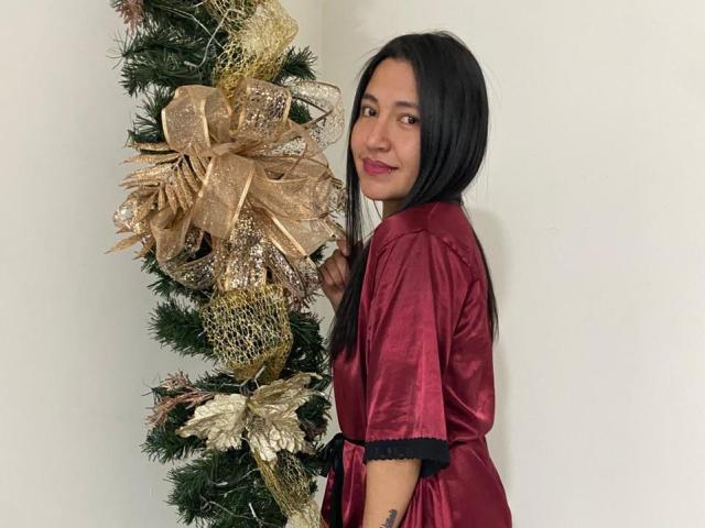 ValentinaHottest with long black hair, wearing a shiny red satin robe, stands beside a decorated Christmas tree with gold ribbons. She's smiling, looking over her shoulder.