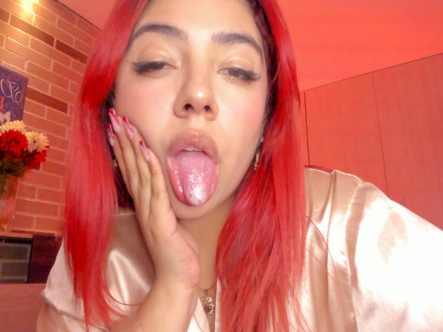 RousseColliinss has red hair, her tongue is out with a glossy, pinkish center, touching her cheek. She's wearing a shiny, beige top with red nails.