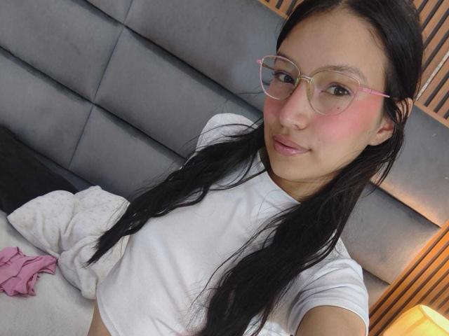SophieDelaney with long black hair, pink-rimmed glasses, blushing pink cheeks, white t-shirt, laying on bed, pink underwear on left, grey upholstered headboard, smiling.