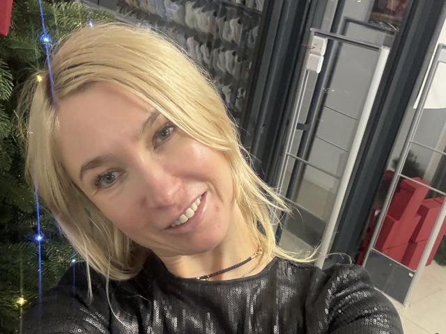 Close-up of WalterCami, a blonde woman with straight hair, smiling at the camera. She has light skin, subtle makeup, and wears a black, textured, long-sleeve top. A thin black necklace is visible. To the left, a decorated Christmas tree with blue lights. The background includes a storage area with shelves and a metal rack. The image is sharply focused on WalterCami's face and upper body.