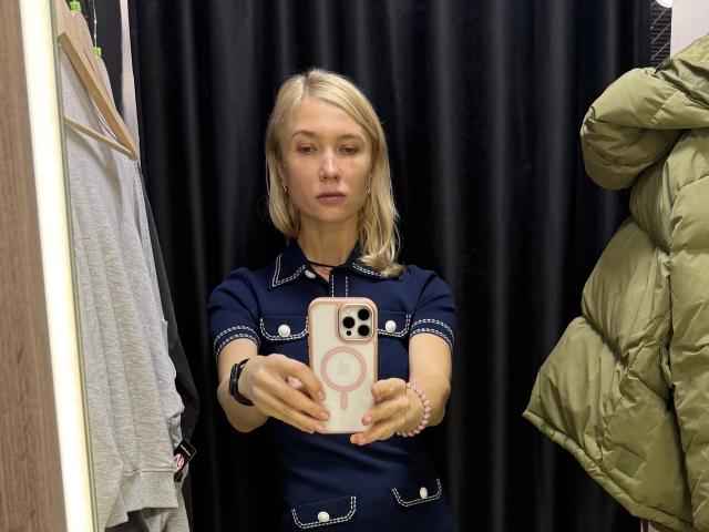 WalterCami stands in a fitting room, taking a selfie with a pink iPhone. She has blonde hair, fair skin, and a neutral expression. She wears a navy blue dress with white embroidered details. Her right wrist has a pink bracelet, and she holds the phone with her right hand. A gray shirt hangs on a wooden hanger to the left, and a green puffer jacket is on a hook to the right. The black curtain background is plain. The image is clear and focused on WalterCami.