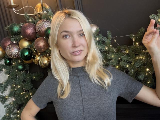 WalterCami, avec des cheveux blonds, portant une chemise grise, tenant un ornement de Noël, debout devant un sapin de Noël décoré avec des ornements colorés et des lumières scintillantes..
