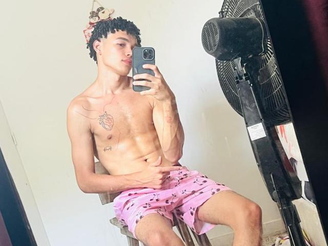 ErikHt, shirtless, with pink shorts, holding phone, sitting on chair, black fan behind, tattoos on chest, curly hair, mirror selfie, arm across belly, looking at phone.