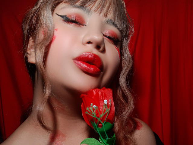 BelleVivienneHt with red makeup, red lipstick, and a red flower near her face, eyes closed, wavy blonde hair, red curtain background, soft light, slightly parted lips.