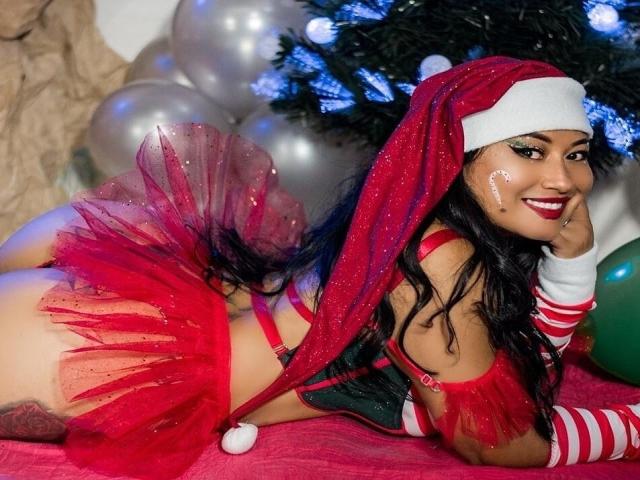 KimoraKim, usando um gorro de Papai Noel vermelho e uma saia de tule vermelha brilhante, está deitada em um cobertor vermelho, mostrando suas nádegas, com luvas listradas e glitter no rosto, sorrindo..