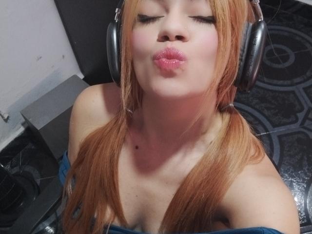 Close-up photo of KataBlonde with light skin and long orange hair in pigtails. She's wearing silver headphones and kissing with shiny pink lips. Her eyes are closed, and she has a small black mole on her right chest. She's dressed in a blue off-shoulder top, showing her upper chest and collarbones. The background is dark and patterned. Her expression is playful and she's centered in the frame.