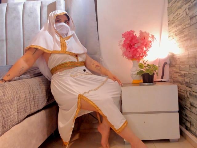 YaraSajar in a white and gold dress, white veil, face mask, sitting on bed, bare feet, tattoos, holding pink flowers on white nightstand, gray bed, stone wall.