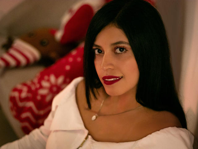 YinelaOslenX with dark straight hair, red lipstick, white off-shoulder top, and small necklace, smiling at camera. Boob outline visible under top. Soft, warm lighting.