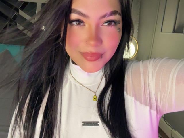 TessaMagic, com cabelos longos e pretos, batom rosa e um piercing no nariz, vestindo uma blusa branca transparente, um colar dourado e uma pequena etiqueta preta com o nome, sorrindo para a câmera..