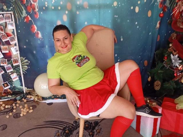 LadySelene with big ass, wearing a green shirt, red skirt, and red knee socks, sitting in a chair, smiling, Christmas tree and presents in the background.