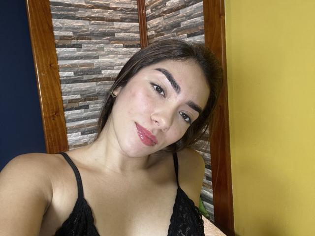 SharonKlart tiene el cabello castaño oscuro, un sujetador de encaje negro y un maquillaje sutil. Está sonriendo, con labios rosados, ojos marrones y senos pequeños. En el fondo, hay paredes de madera y de color amarillo..