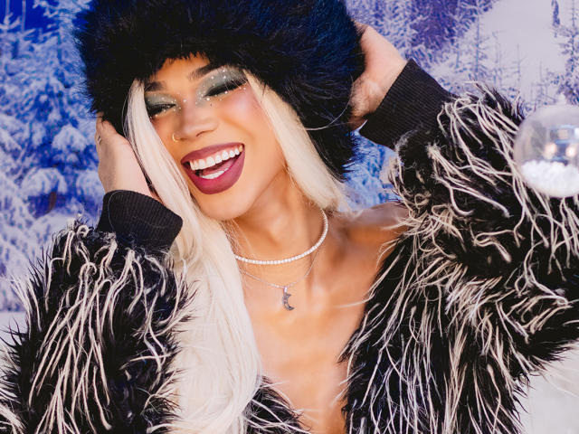 NaomiSteward with blonde hair, black fur hat, and fluffy black-and-white jacket, smiling with red lipstick, green eyeshadow, and hands in hat, wearing a diamond necklace.