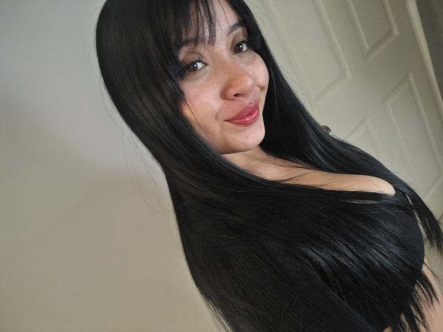 LunaBellaHotty with long black hair, smiling at the camera, showing her tits in a low-cut top, looking cute and sexy with red lips.