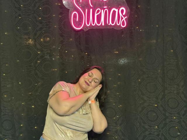 LadySelene with closed eyes, hands near face, smiling, wearing a shiny beige top, standing in front of a pink neon sign, dark patterned background, slight red neon light.