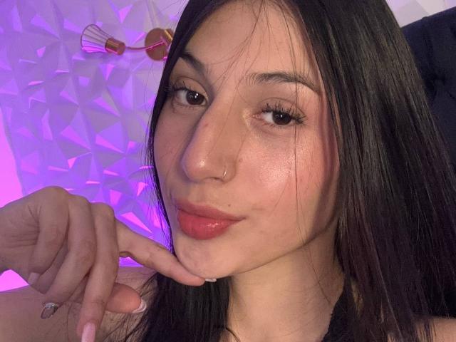 SophieBeaumont has long dark hair, a nose ring, and is slightly smirking with pink lipstick. Her hand's finger is near her chin. She's looking at the camera.