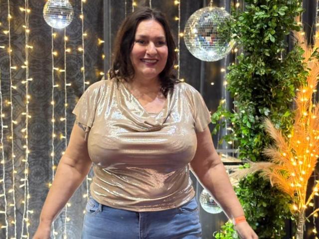 LadySelene with large tits, wearing a shiny, gold, cowl-neck top, and blue jeans, standing in front of sparkling lights and mirrored balls, smiling, with brown hair.
