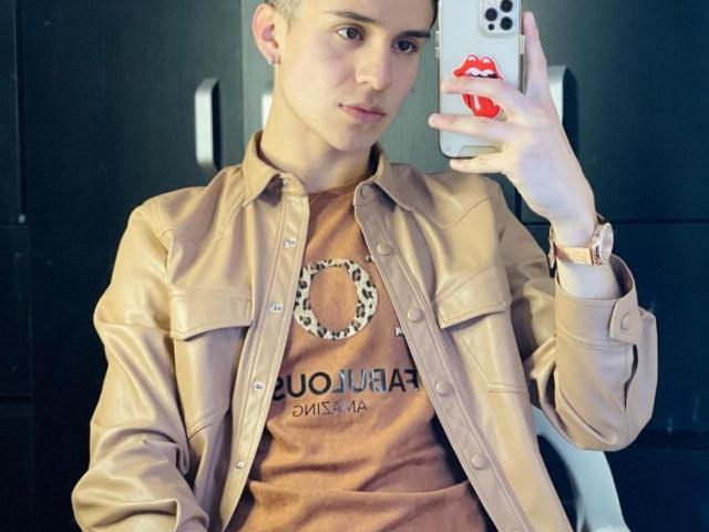 JonyGumm with short hair, holding phone with red lip sticker, wearing tan leather jacket over brown shirt, pink watch, sitting, taking selfie, looking at camera, slightly smiling.