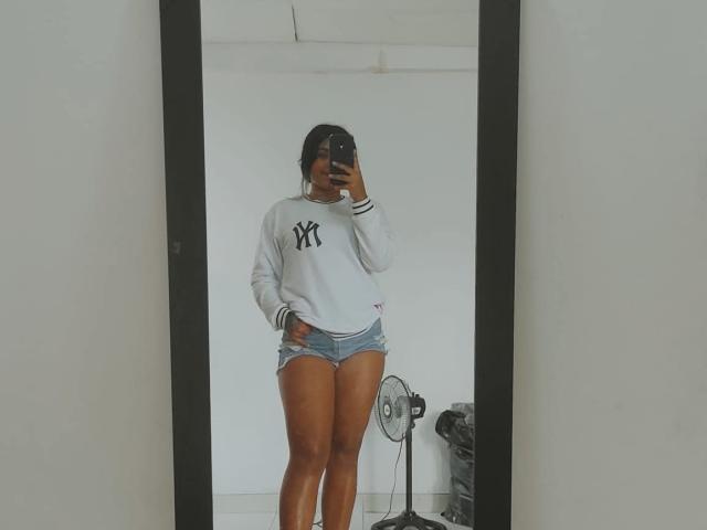 CurvyBrunnete in a white NY Yankees sweater and blue denim shorts, standing in front of a mirror, holding a phone, with a fan and chair in the background.