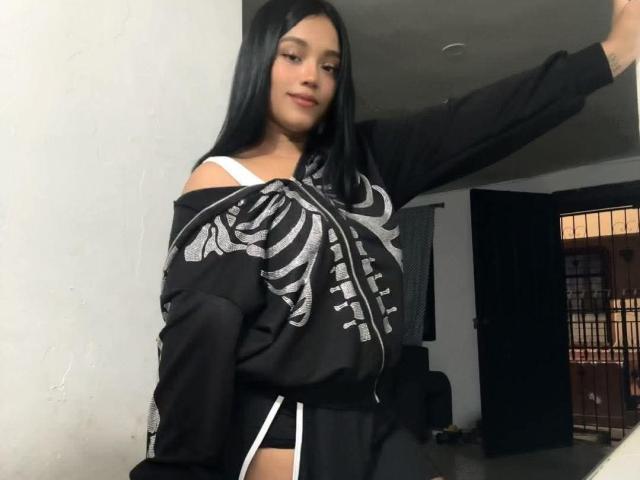 CurvyBrunnete with long black hair, black skeleton shirt, and white underwear, has one arm raised, showing off her small tits and a slight smile.