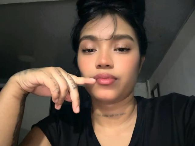 CurvyBrunnete with dark hair in a messy bun, pouting lips, and finger touching them, wearing a black shirt, has a tattoo on her finger and a necklace.
