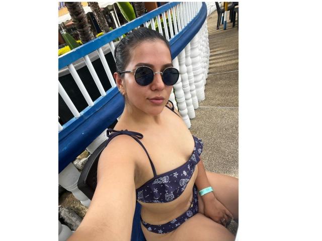 Velka with dark skin, black sunglasses, and a blue patterned bikini, showing her small tits and sitting with her legs apart, wearing a green wristband.