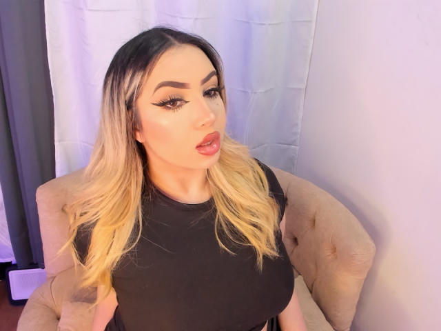 Melissa Lust, avec ses cheveux blonds dégradés, un haut noir et du maquillage, faisant une moue avec les lèvres légèrement entrouvertes, assise sur une chaise beige capitonnée, montrant sa poitrine généreuse..