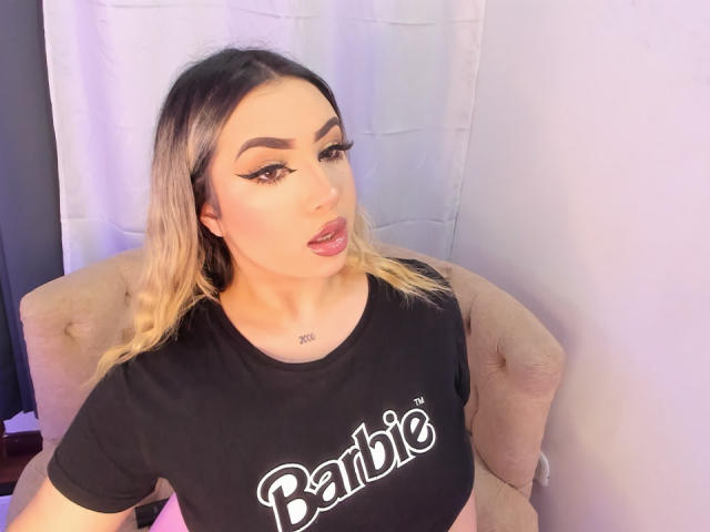 MelissaLust with blonde hair, bold eyebrows, and winged eyeliner, wearing a black "Barbie" shirt, has her mouth slightly open, showing her pink lipstick and small tattoo on her neck.