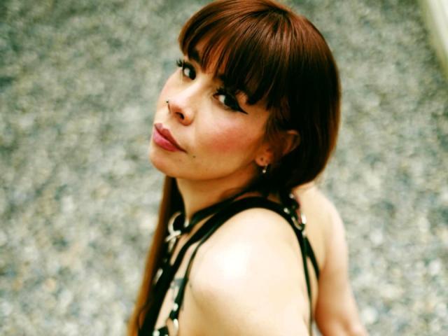 AriiannaHot with brown hair and bangs, nose ring, and pink lipstick, looking back over her shoulder, wearing a black and white halter top, showing her bare back.