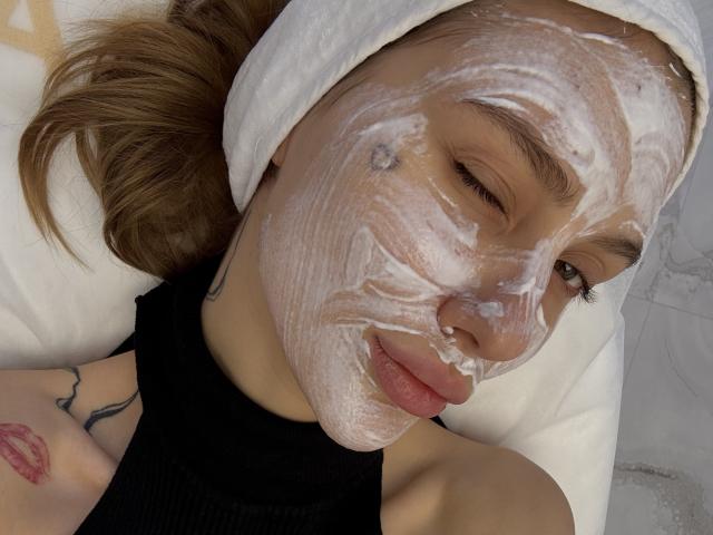 Close-up of SensualMila with a towel on her head, lying down. Her face is covered in thick white facial cream, with one eye slightly closed. She has light brown hair, a small nose piercing, and a tattoo on her right shoulder of pink lips. She's wearing a black sleeveless top. Her eyebrows are thin and dark, and she has long eyelashes. The texture of the facial cream is smooth and slightly glossy. The background is mostly white and blurred. SensualMila's expression is relaxed. The image focuses on her facial cream application.