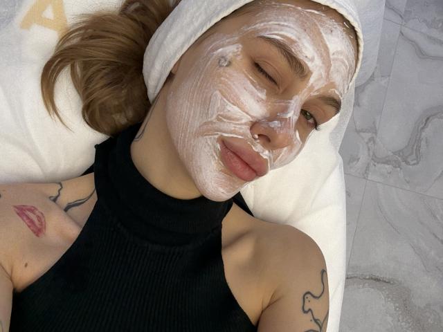 SensualMila has a white towel on her head, with thick face mask covering her eyes and nose. She's wearing a black sleeveless top, lying down with eyes closed.