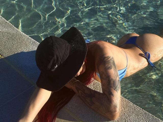 SofiaLeenss lying face-down in a pool, wearing a black hat and blue thong bikini, with tattooed arms and red hair, showing her ass and small, round butt.
