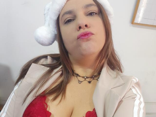 AmeliaWilson with brown hair, wearing a white Santa hat, pink lipstick, red lace bra, beige jacket, and black necklace, pouting slightly. Boobs visible. 

 