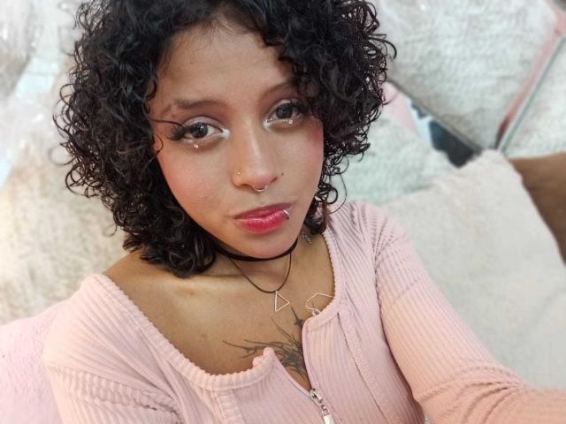 LunaKeller with curly black hair, pink ribbed top, nose ring, and pink lipstick, looking up with eyeliner and subtle makeup. Her small tits are partially visible.