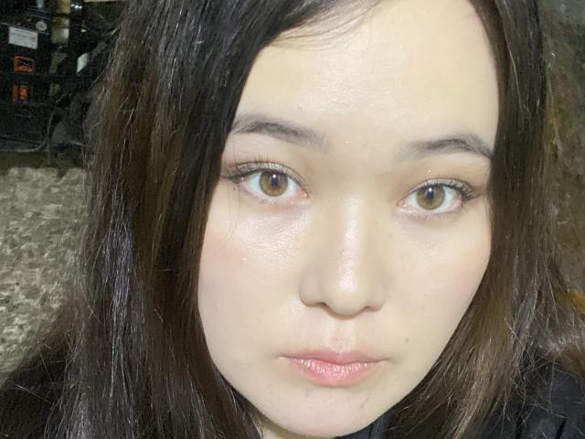 LiyaFlower's face with pale skin, brown eyes, and pinkish lips, wearing a black top, has straight brown hair, and a slightly shiny forehead. She looks straight at the camera.