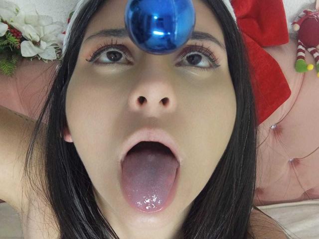 JeneKurz with black hair, blue ball on forehead, mouth open, tongue out, drooling, eyes looking up, wearing Santa hat, pink cushioned background.