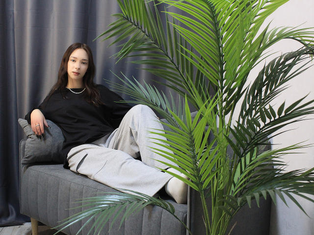 CiliHot with long brown hair, black shirt, and grey pants, lounging on grey couch, next to large green palm, looking relaxed, wearing white socks, ring on right hand.