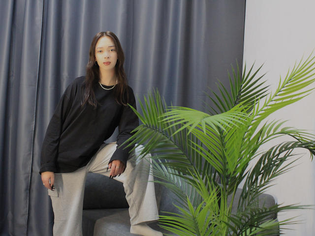 CiliHot with long brown hair, wearing a black sweater and beige pants, sits on a gray chair, hands on knees, next to a green palm plant.