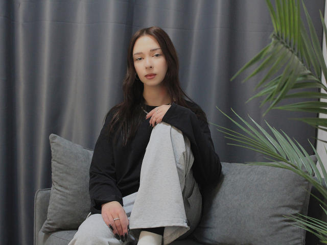 CiliHot with long brown hair, black top, and gray pants, sitting on a gray couch, holding a gray blanket, next to a green plant, looking calm.