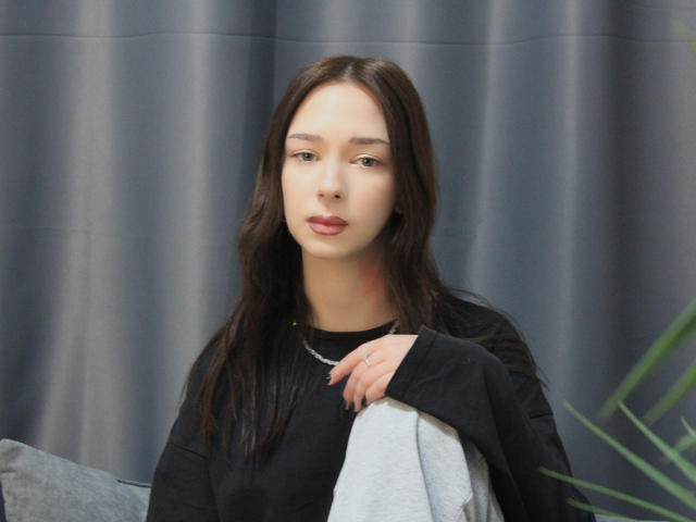 CiliHot with long brown hair, pale skin, and pink lips, wearing a black shirt, holding a gray blanket, gray curtains in background, subtle green necklace.