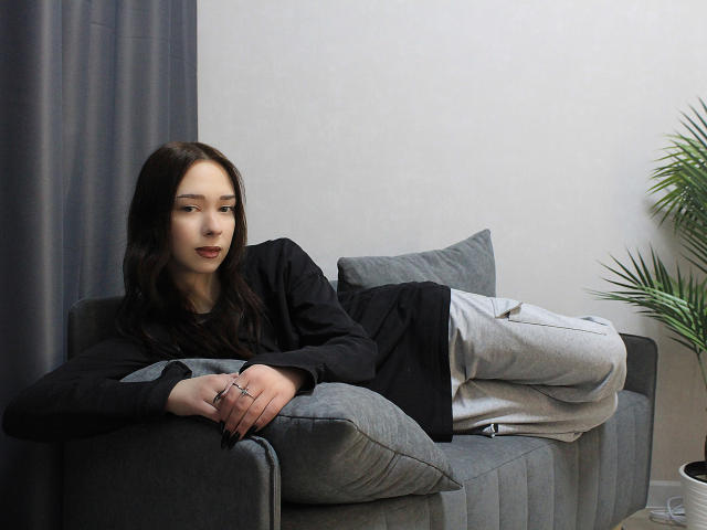 CiliHot with long dark hair, wearing black shirt and gray pants, lounging on gray couch, arms resting on armrest, fingers with ring, looking slightly bored, potted plant right.