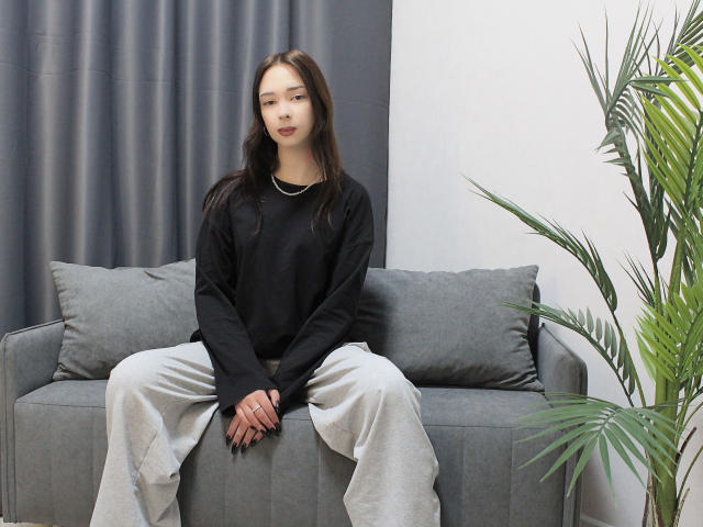 CiliHot with straight brown hair, black shirt, and gray pants sits on a gray couch, hands on knees, looking slightly bored. A green plant is on the right.