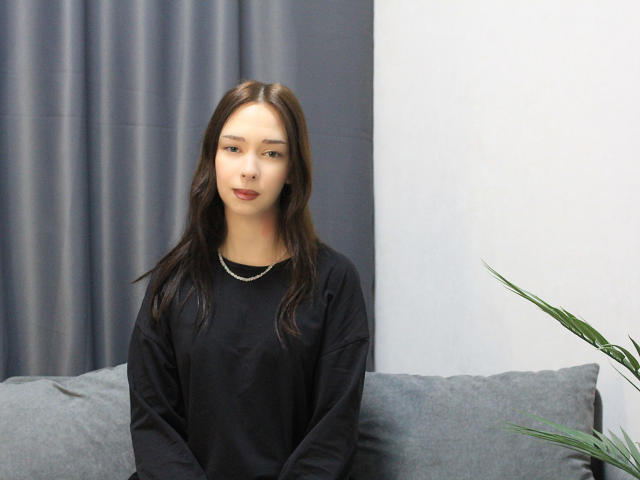 CiliHot with medium tits, brown eyes, and long brown hair, wearing a black shirt, sitting on a grey couch, looking at the camera with a neutral expression.