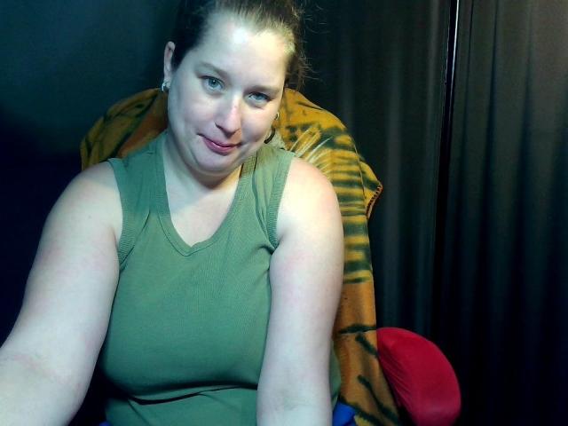 Roosje88 with pale skin, brown hair in a bun, green sleeveless top, sitting in a chair with a tiger-patterned blanket, red seat, smiling slightly. Boobs visible under the top.