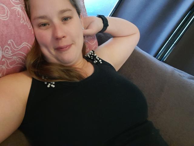 Roosje88 is a chubby woman with light skin, brown hair, and small tits, wearing a black sleeveless top with white bead details, lying on a pink pillow.
