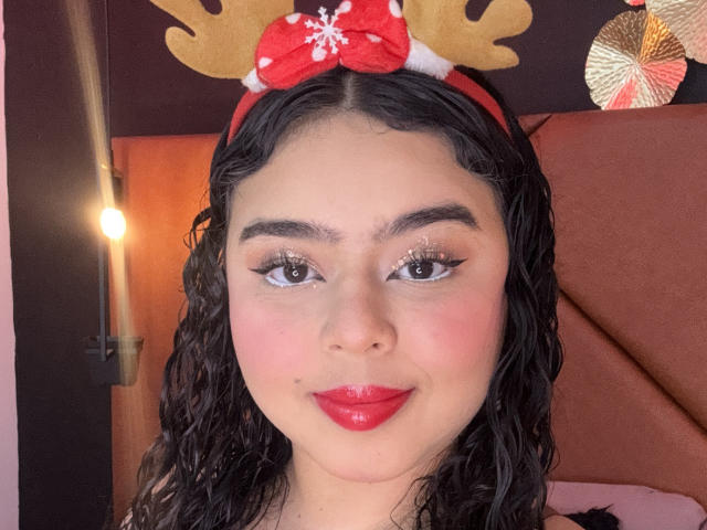 AprielKeith with curly black hair, wearing a red bow with white snowflakes, red lipstick, and winged eyeliner, smiling at the camera, in a headshot with warm lighting.