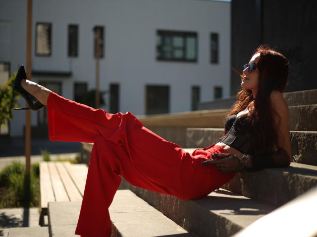 OliviaMuur is lounging on stone steps, wearing red pants, black high heels, and a black lace top, with sunglasses on, her legs up, in bright sunlight.