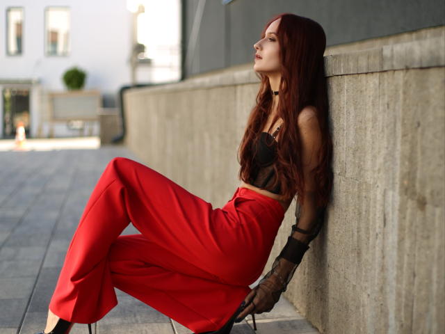 OliviaMuur with red hair, black sheer top, and red pants, leaning against a concrete wall, wearing black mesh gloves, legs crossed, looking to the side, in sunlight.