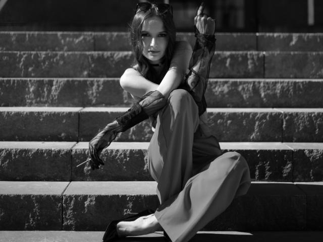 Black-and-white photo of OliviaMuur sitting on stone steps. She's wearing a sleeveless top, wide pants, and black flats. OliviaMuur's right arm is wrapped in a sheer, black, floral-patterned sleeve. She's holding a cigarette in her right hand, resting on her knee. Her left arm is crossed over her right. OliviaMuur has wavy hair, sunglasses on her head, and a serious expression. High contrast, shadows, and textures of the stone steps are prominent. Her legs are bent, and she's positioned slightly to the right in the frame.